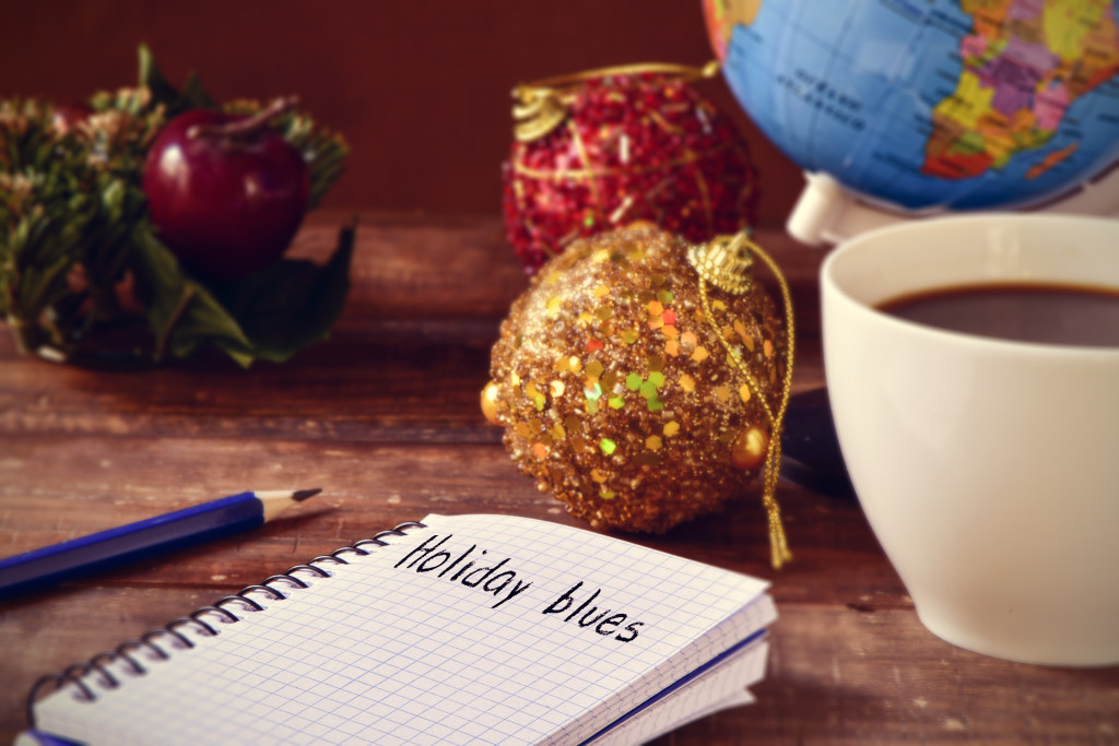 closeup of a notebook with the text holiday blues written in it on a rustic wooden table with some christmas ornaments, a cup of coffee and a globe